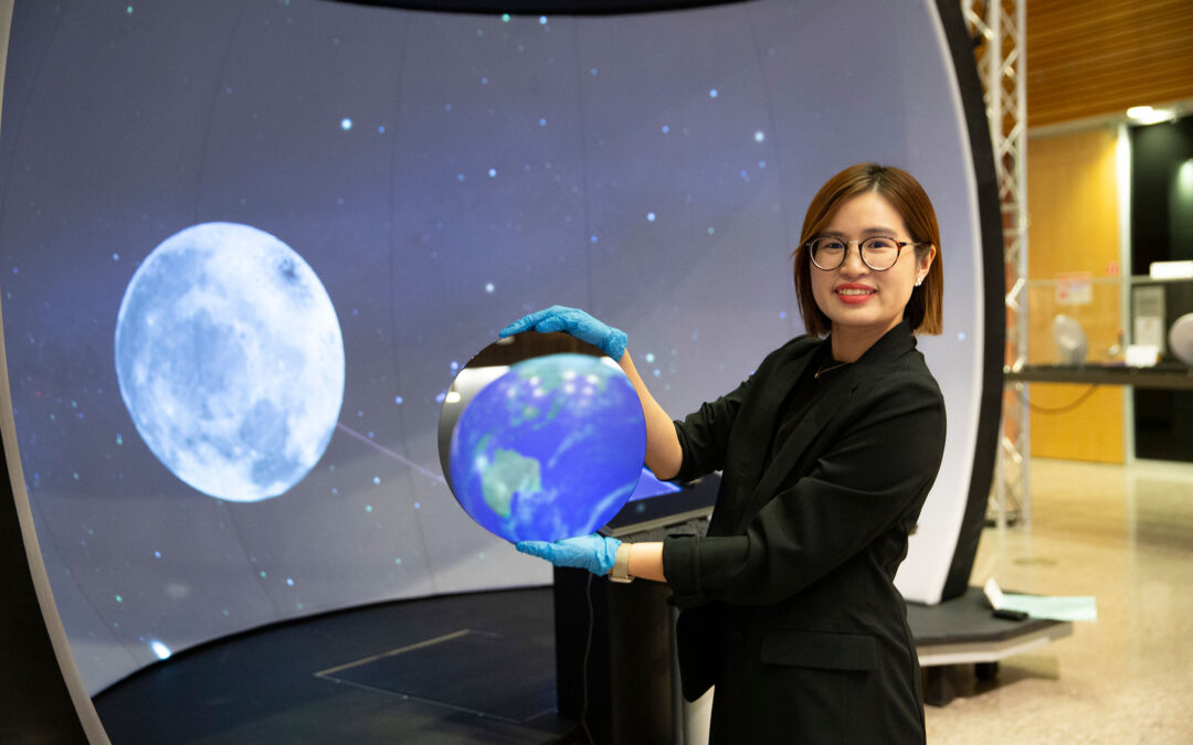 Ying-Chen "Daphne" Chen holds semiconductor material reflecting a model of the Earth in front of a screen showing the moon