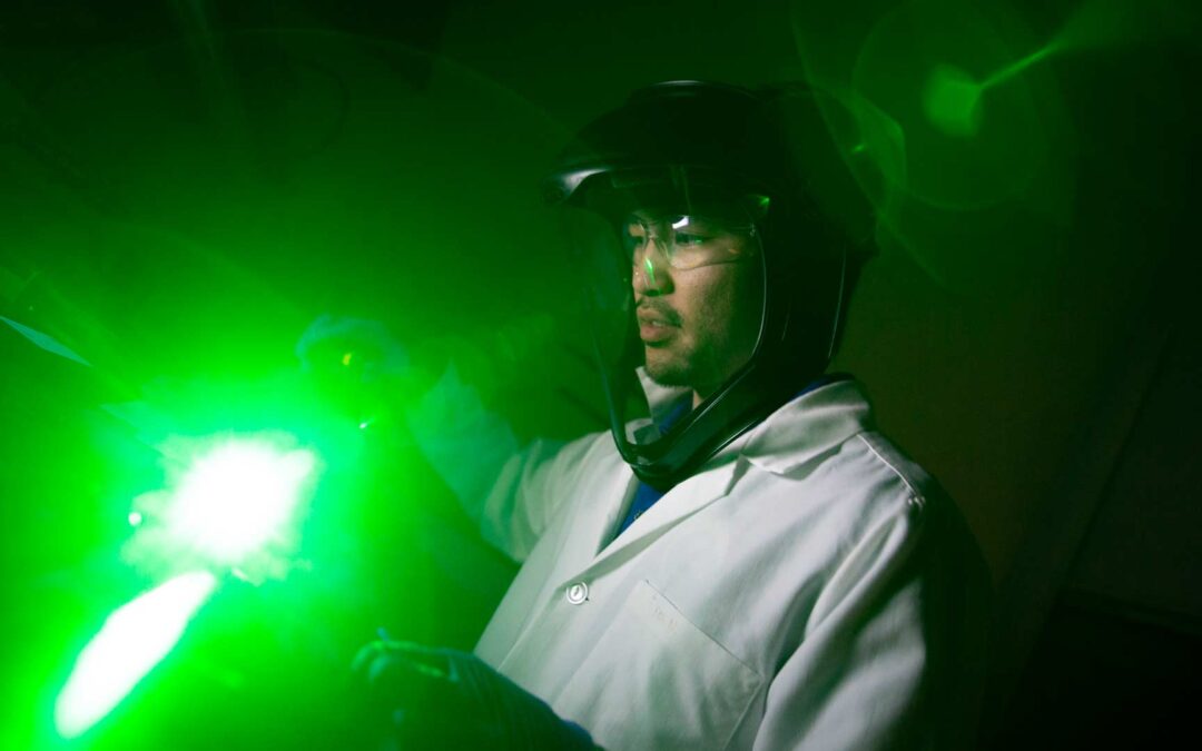 A man wearing protective equipment works with lab devices giving off a bright green glow.