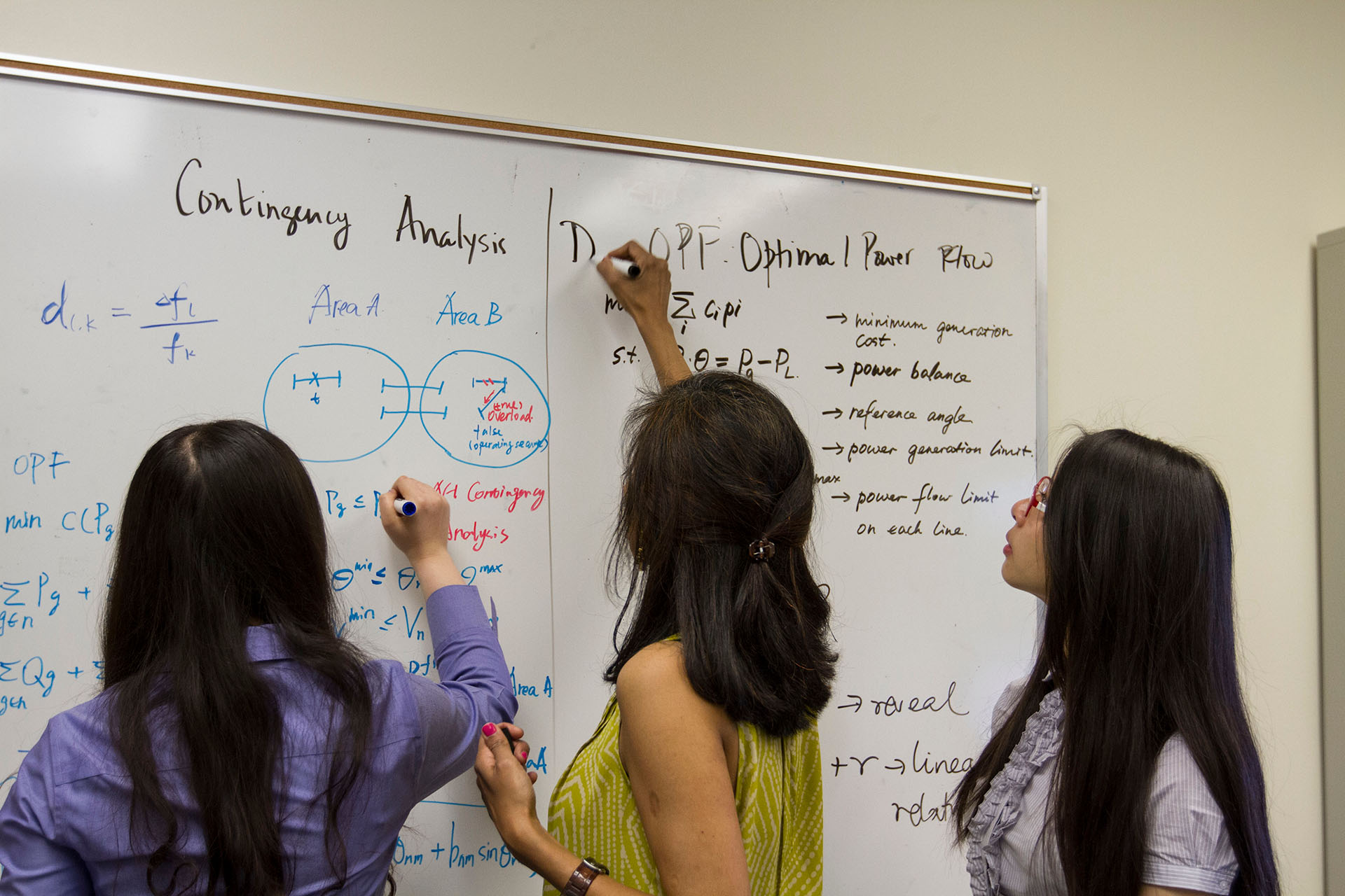 Three women draw on a whiteboard filled with equations and diagrams