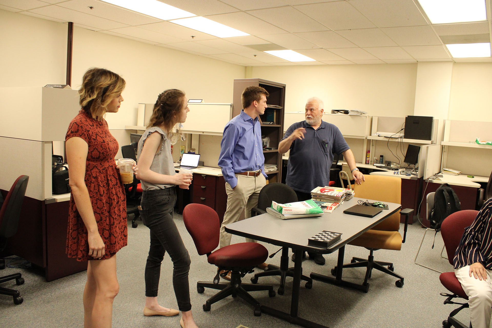 Three people look on as another speaks while gesturing behind a table in a room