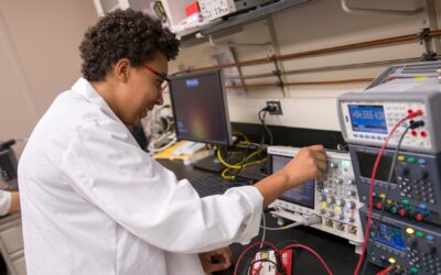 A person in a white lab coat is operating electronic test equipment, adjusting settings on one of the electrical devises visible in the frame.