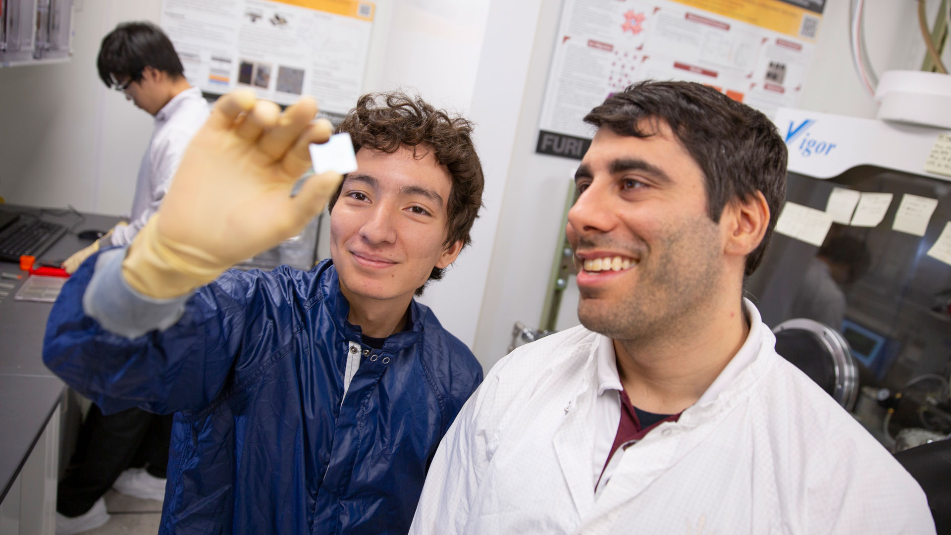 An ASU professor and student stand side by side in a lab. The student holds a small object between his thumb and forefinger that they both look at, smiling.